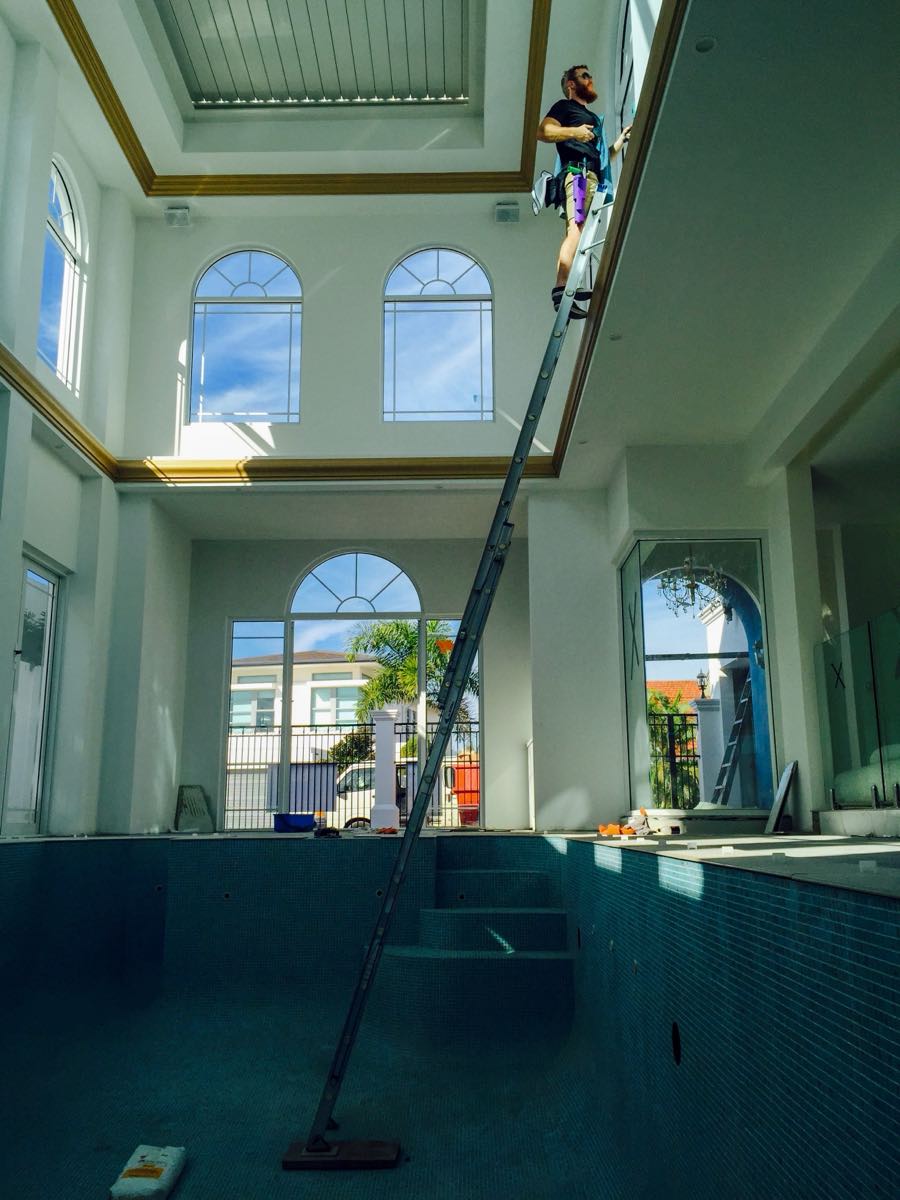 Rhys up a ladder cleaning high arched interior windows above an empty indoor pool in a large Gold Coast home, Streakers era